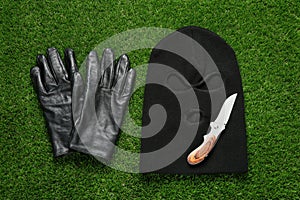 Black knitted balaclava, gloves and knife on green grass, flat lay