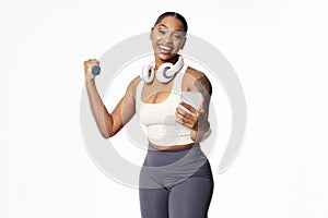 black lady holds smartphone flexing arm muscles with dumbbell, studio