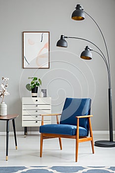 Black lamp next to blue wooden armchair in grey apartment interior with poster and plant. Real photo