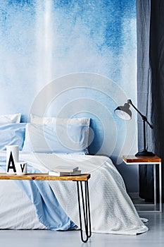 Black lamp on wooden table next to a white bed against blue wall