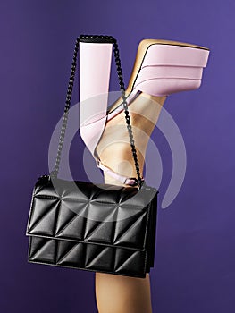 Black leather bag hangs on high heel of pink shoe put on leg