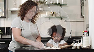 Black little girl with her mother making dough in the bright kitchen - covering the dough with flour