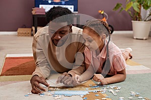 Black little girl solving jigsaw puzzle laying on floor with father