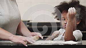 Black little smiling girl with her mother rolling out the dough in the bright kitchen