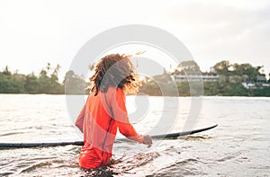 Black long-haired teen boy with a surfboard ready for surfing. He walking into Indian ocean waves with magic sunset background.