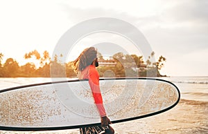 Black long-haired teen boy with a waxed surfboard ready for surfing with sunset backlight. He walking into Indian ocean waves.