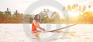 Black long-haired teen man floating on long surfboard, waiting for a wave ready for surfing with palm grove litted sunset rays.