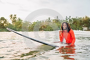 Black long-haired teen man showing shaka sign floating on long surfboard, waiting wave ready surfing with palms litted sunset rays