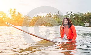 Black long-haired teen man showing shaka sign floating on long surfboard, waiting wave ready surfing with palms litted sunset rays