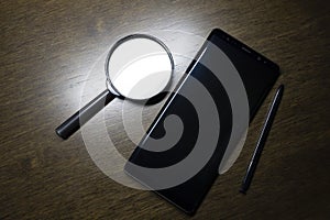 A black magnifying glass with a black laptop and a black pen are on the brown wooden working table