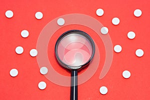 Black magnifying glass is on red background surrounded by white pills as ornament polka dots. Medical or pharmaceutical concept by