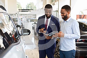 Black Male Customer In Car Dealership Center Looking At Brochure With Salesman