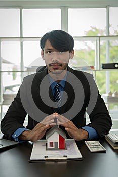 Black male real estate agent puts a miniature model of a house on a table inside a real estate sales office.