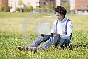 Black man with afro hair using his laptop sitting on skateboard on the grass of an urban park.