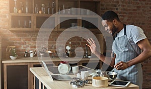 Black man baking pastry and using laptop in kitchen
