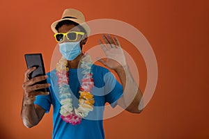 Black man in carnival costume and pandemic mask holding  a mobile phone isolated on orange background. African man in various