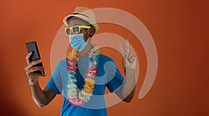 Black man in carnival costume and pandemic mask holding  a mobile phone isolated on orange background. African man in various