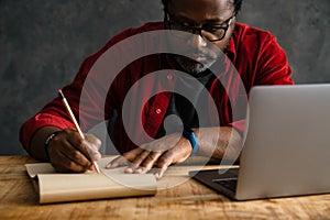 Black man in eyeglasses writing down notes while working with laptop