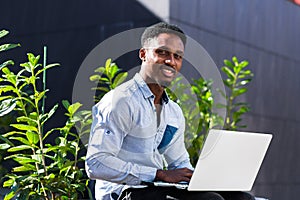 Black man freelancer working online using laptop sitting on bench outside office modern building in city urban park on street.