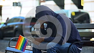 Black man in hood sitting on bench with lgbt minority flag, preconceptions