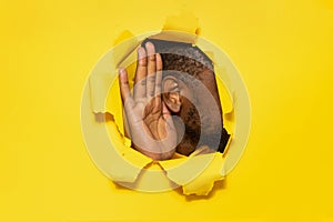 Black man& x27;s ear and hand through a torn hole in yellow paper background, free copy space, closeup