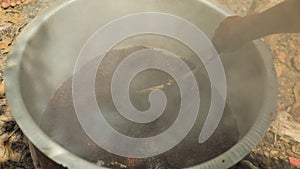 Black man's hand is roasting some seeds in huge aluminium pot with heavy smoke.