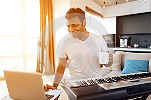 A black man sits in the living room of his apartment and plays a synthesizer. He composes music and drinks coffee.