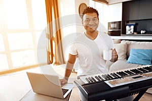 A black man sits in the living room of his apartment and plays a synthesizer. He composes music and drinks coffee.