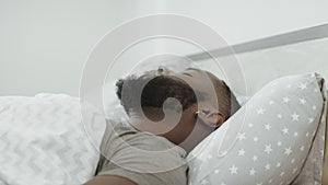 Black man sleeping in bed in morning. Young adult covering head with pillow.