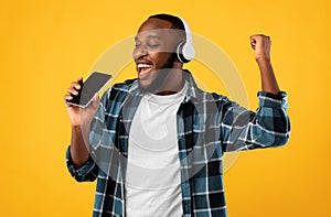 Black Man Wearing Headphones Singing Holding Smartphone Over Yellow Background