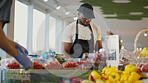 A Black man in a white T-shirt and a black apron in a black cap sorts vegetables on the counter in a modern grocery