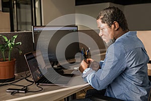 Black Man Writing Lines of Code On Desktop PC With Two Monitors and a Laptop Aside in Stylish Office. Professional Male