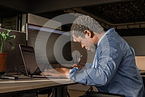 Black Man Writing Lines of Code On Desktop PC With Two Monitors and a Laptop Aside in Stylish Office. Professional Male