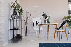 Black mannequin`s leg on wooden chair in elegant dining room interior with copy space on the empty wall, flowers and leaf in vase