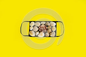 Black medical face mask and seashells on a yellow background. Flat lying, copy space, holiday cancellation, pandemic, tourism