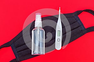 Black medical protective face mask with hands sanitizer and digital thermometer on red background. Protection against virus, flu