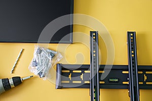 Black metal bracket for wall mounting a computer monitor or TV, screwdriver, mounts and monitor screen on a yellow background. The