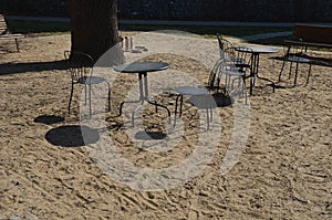 Black metal chairs and round tables in a cafe on a cobbled square with a sand surface of compacted yellow gravel under an old tree