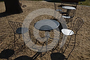Black metal chairs and round tables in a cafe on a cobbled square with a sand surface of compacted yellow gravel under an old tree