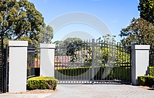 Black metal driveway entrance gates set in brick fence with garden trees in background