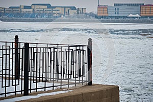 Black metal fence of the city embankment. The Amur river during ice drift and the Chinese coast in a blurred background.