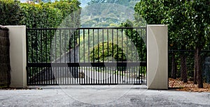 Black metal wrought iron driveway property entrance gates set in brick fence, concrete path, garden trees