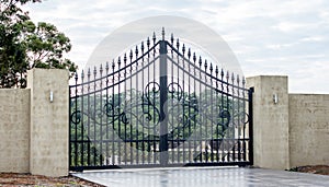 Black metal wrought iron driveway property entrance gates set in concrete brick fence, lights, garden trees in background