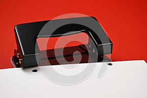 Black metallic hole puncher isolated on red and a paper with two holes.