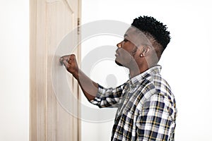 Black Millennial Man Knocking At Closed Door Of Apartment Indoors