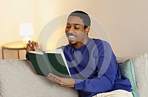 Black millennial man reads paperback book sitting in contemporary home