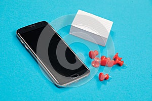 Black mobile phone, a pile of white scratch paper and red push pins are lying on pale-blue surface isolated