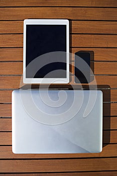 Black mobile phone, tablet, metallic laptop outdoor on an orange wooden table