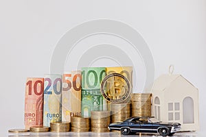 A black model of a car, house and ten, twenty, fifty, one hundred, two hundred and coins euro rolled bills banknotes on white
