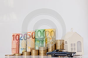 A black model of a car, house and ten, twenty, fifty, one hundred, two hundred and coins euro rolled bills banknotes on white